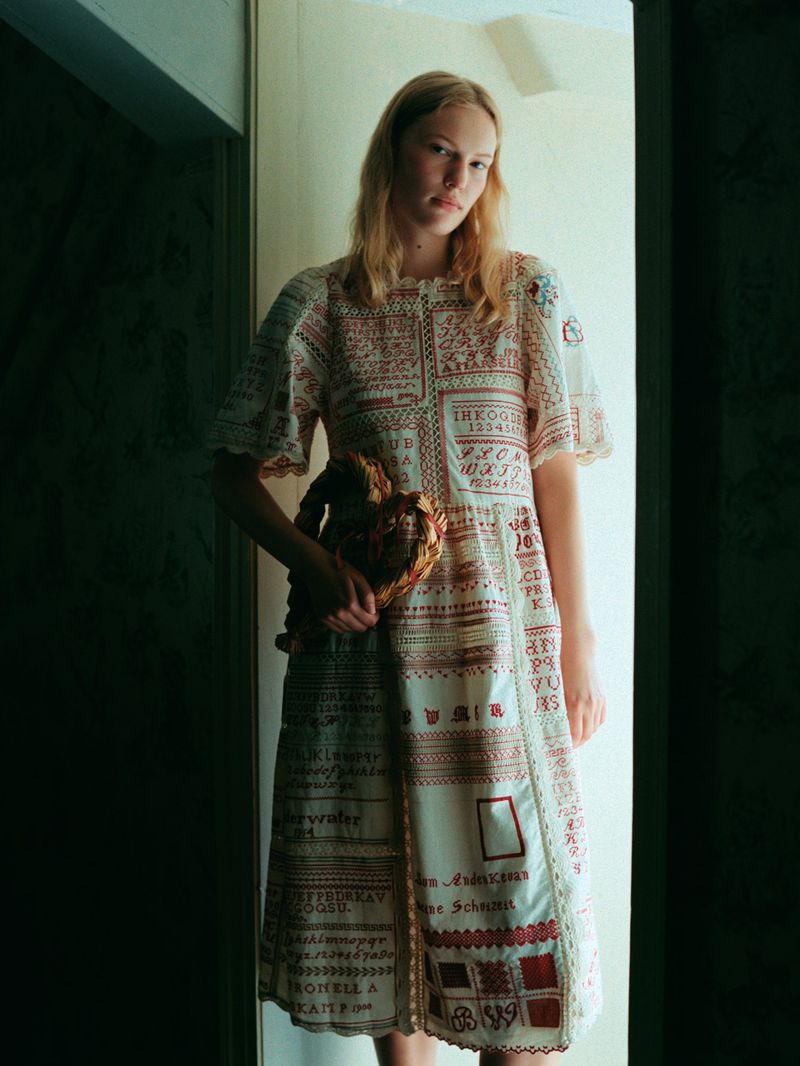A fair-skinned adult female with blonde hair wearing a patchwork dress, holding an organic object, stands indoors in natural light.