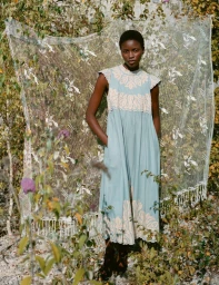Model standing outdoors wearing a light blue embroidered dress in front of a draped lace fabric backdrop