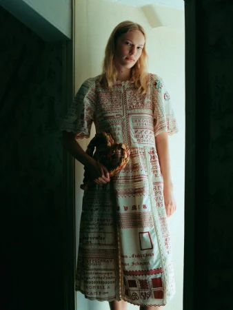 Model wearing patterned embroidered dress holding a woven handbag standing in a doorway