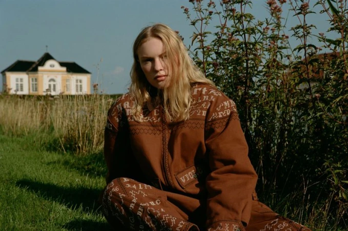 Model wearing a brown patterned jumpsuit seated outdoors in a grassy field with a building and foliage in the background