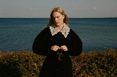 Model in a black coat with an elaborate white lace collar posing outdoors against a backdrop of open water.