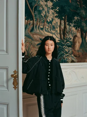 Model standing in an ornate interior with a landscape tapestry backdrop, wearing a dark jacket.