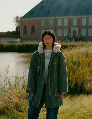 Model standing outdoors wearing an embroidered green parka coat and blue jeans near a body of water and a large brick building