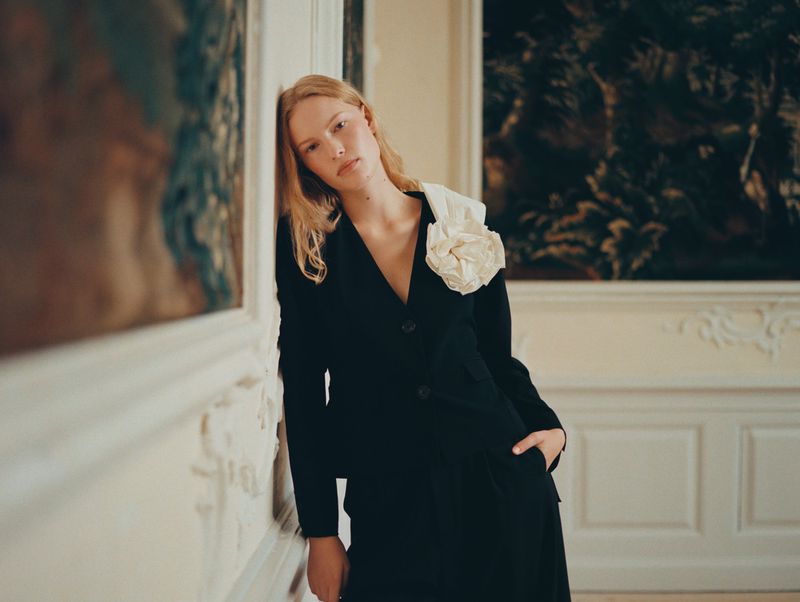 Model leaning against a wall in an interior space wearing a black jacket with a large white flower accent.