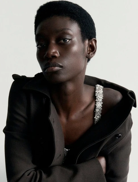 A fashion editorial shot in a photo studio featuring a female model in a green jacket and skirt posing in front of a white backdrop.