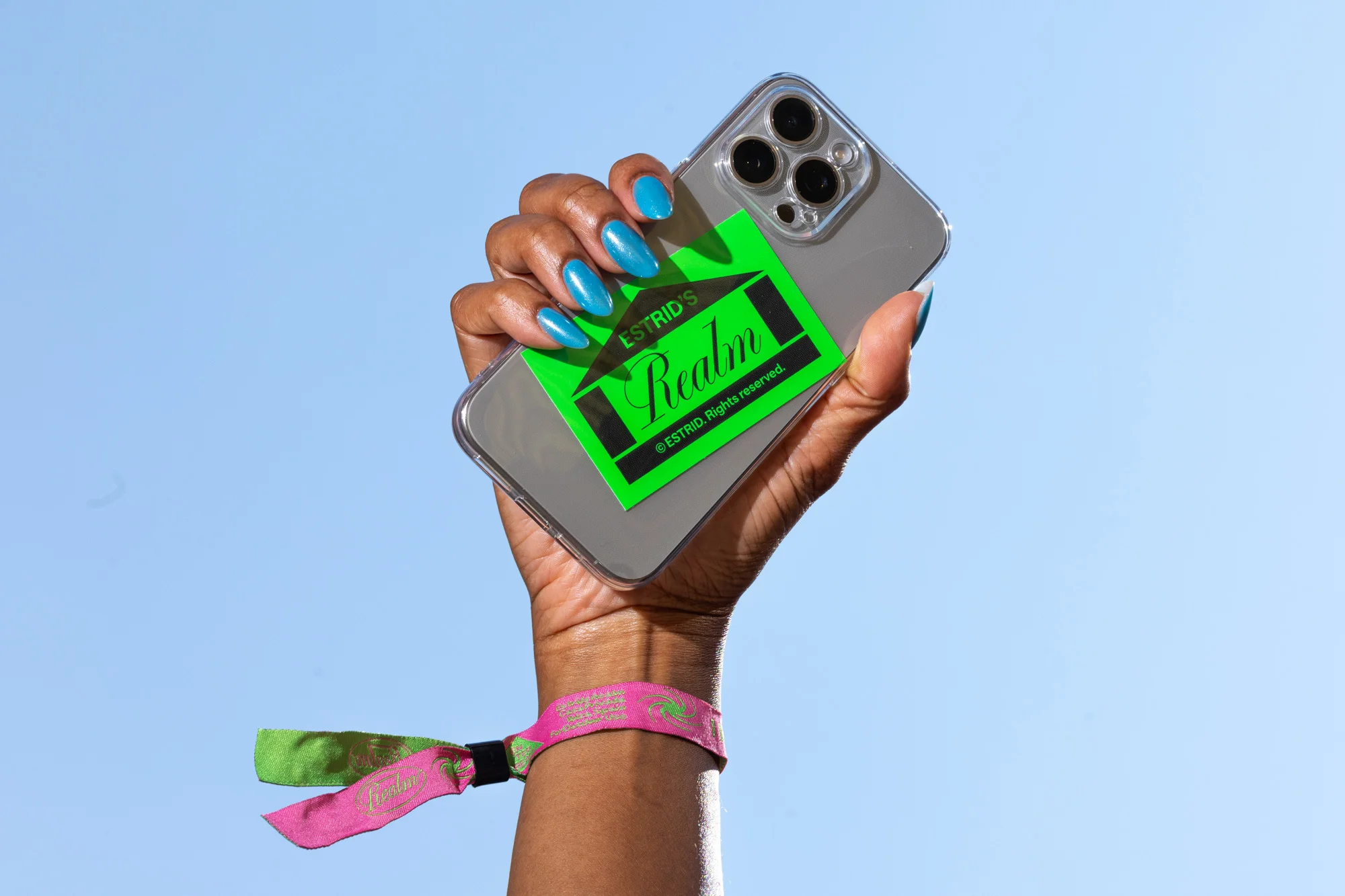 Hand holding an iPhone with a green sticker featuring brand name 'Estrid's Realm', blue nails, against a clear blue sky background.