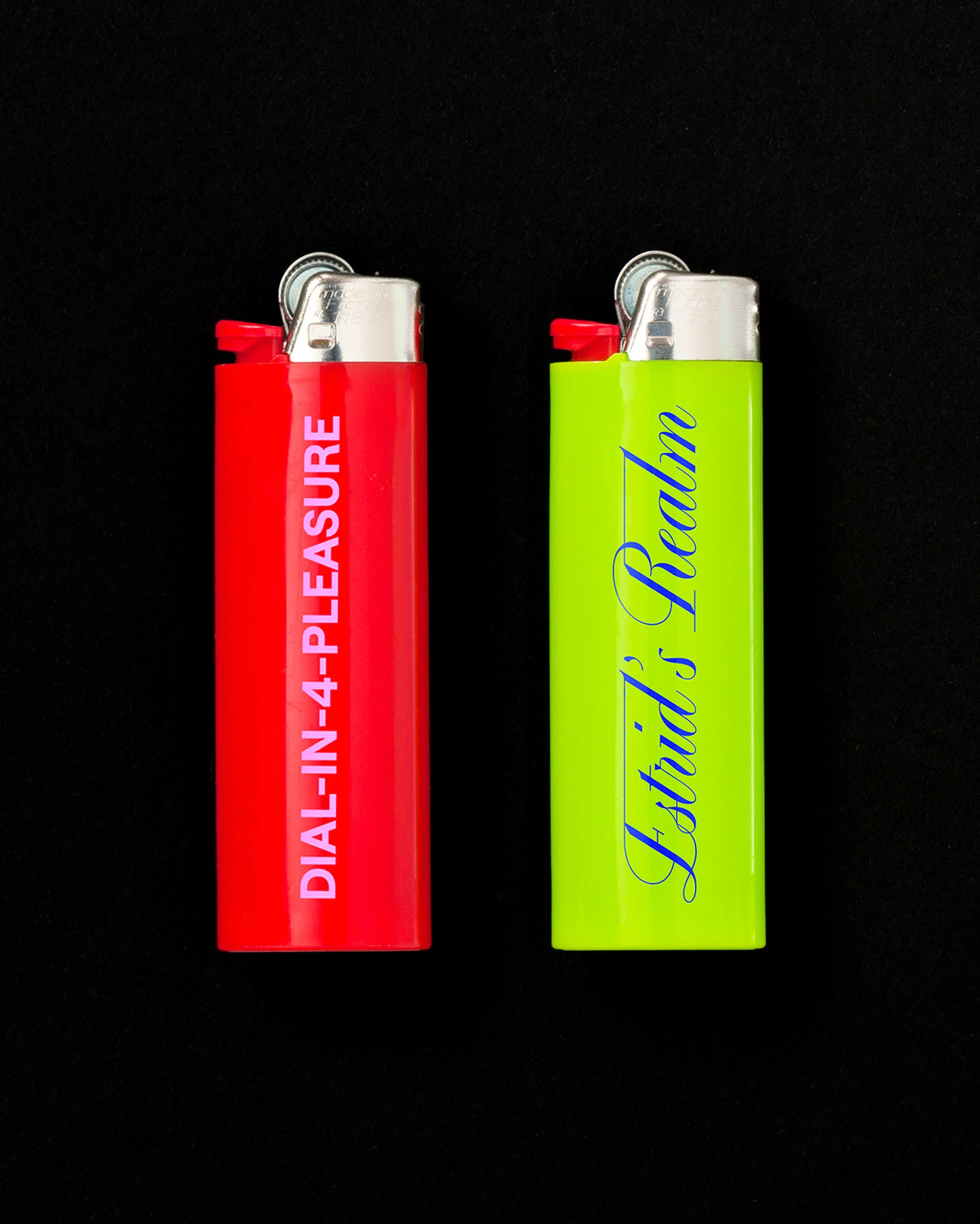 Two lighters, one red with "DIAL-IN-4-PLEASURE" text and one green with "Estrid's Realm" text, are side-by-side on a black background.