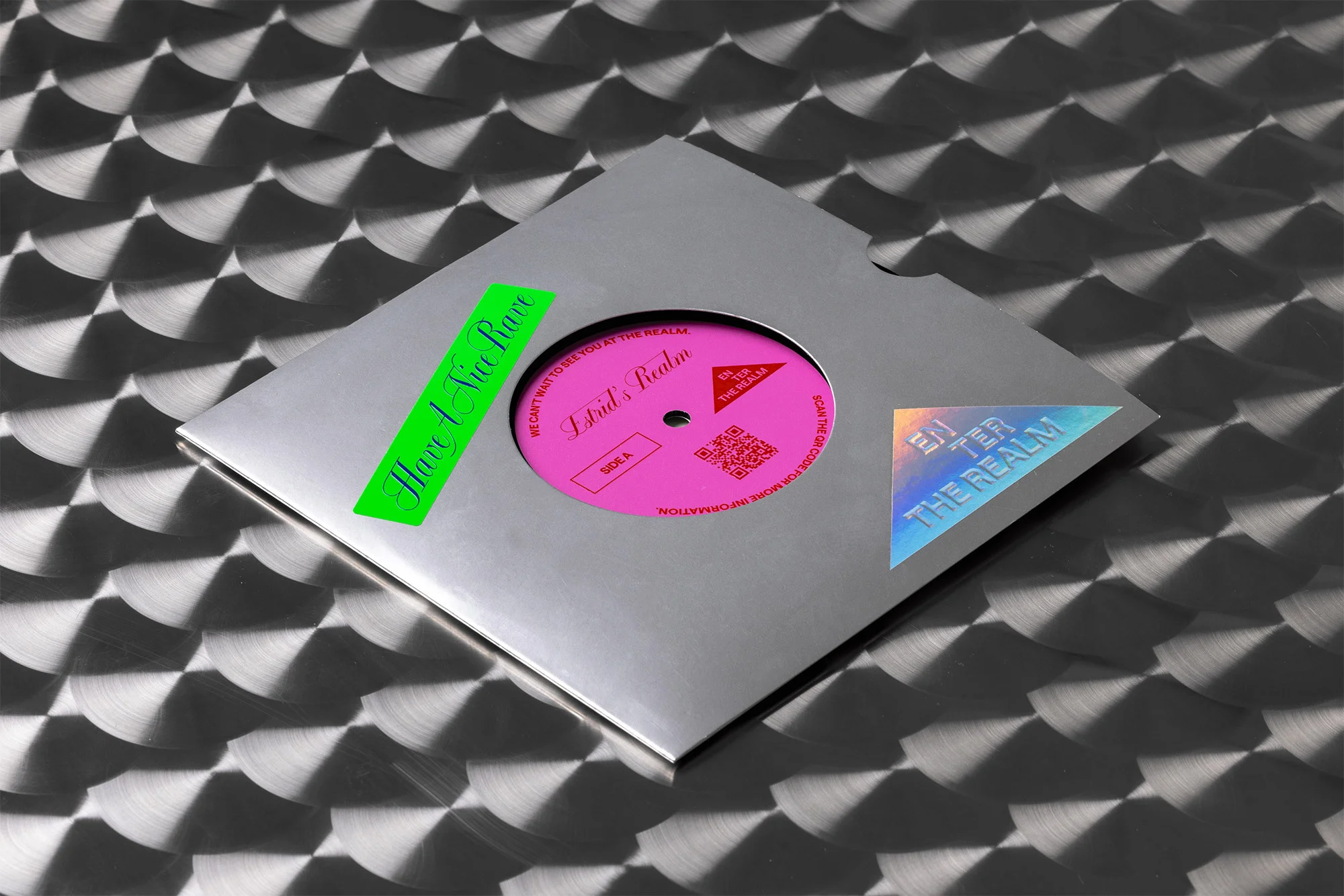Close-up of a pink vinyl record in a silver sleeve with stickers on a textured metallic surface, promoting music.