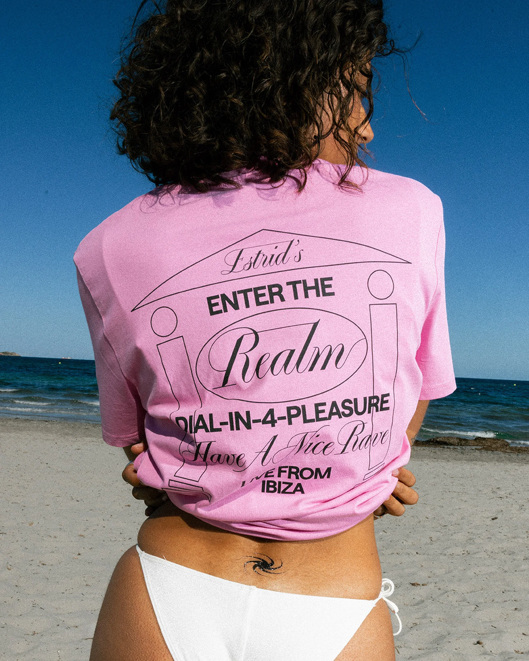 Woman in pink t-shirt with graphic stands on beach against sky. Text includes, Estrid's, Enter the Realm, Dial-in-4-Pleasure, Ibiza.