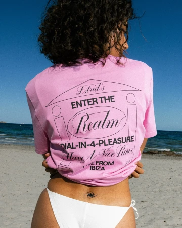Woman in pink t-shirt with graphic stands on beach against sky. Text includes, Estrid's, Enter the Realm, Dial-in-4-Pleasure, Ibiza.