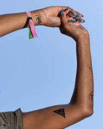 Close-up of arms raised against a clear blue sky, featuring temporary tattoos, turquoise nail polish, and a pink and green wristband.