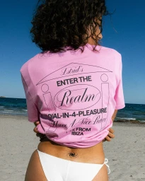 Woman with curly dark hair wearing a pink t-shirt and white bikini bottom standing on a sandy beach