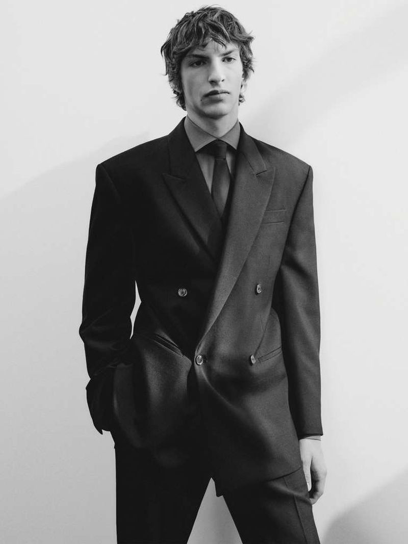 Monochrome portrait of a young adult man in a double-breasted suit with a dark shirt and tie, posing against a plain white wall.