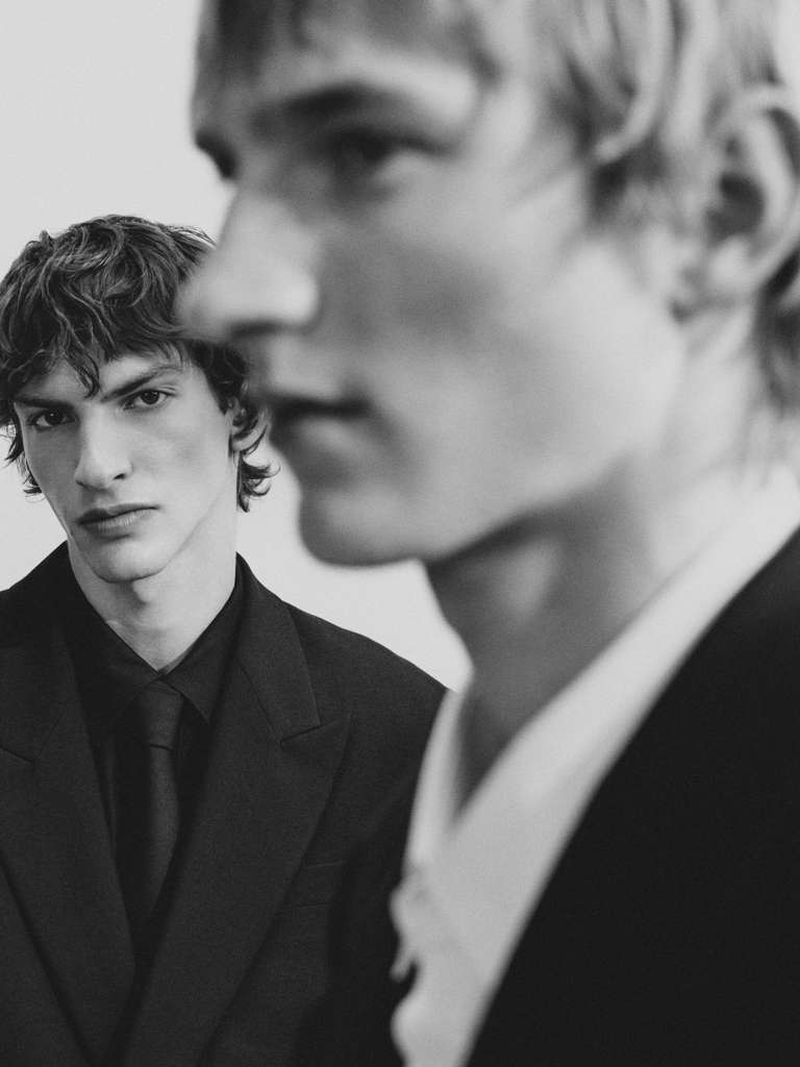 Two men in black suits pose in a studio setting, one with curly hair looking at the camera, the other partially out of focus.