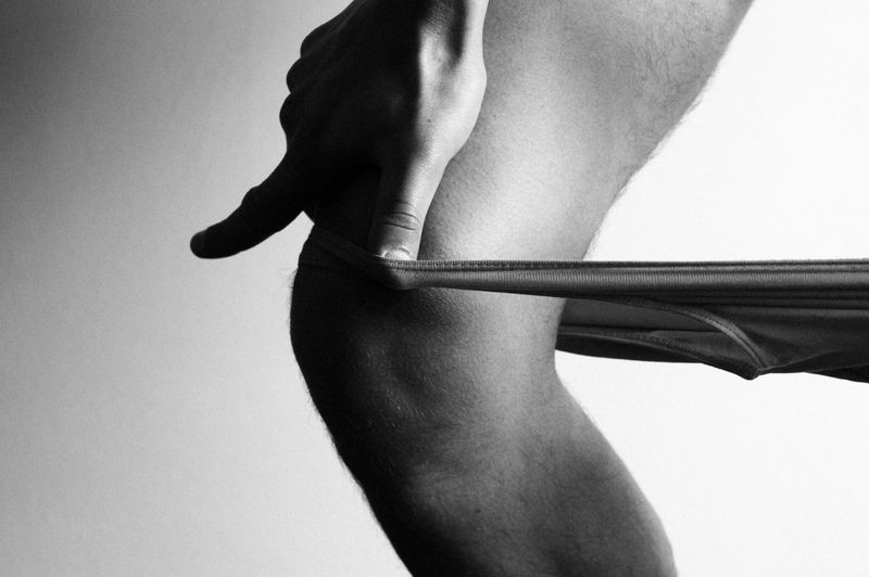 Monochrome image of a person's leg and hand holding underwear, captured in a photo studio against a plain backdrop.