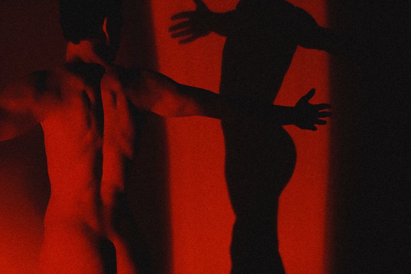 A shirtless man viewed from behind under dramatic red lighting, casting a dark silhouette on a wall for an editorial project.