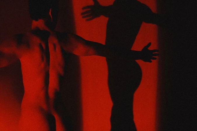 A shirtless man viewed from behind under dramatic red lighting, casting a dark silhouette on a wall for an editorial project.