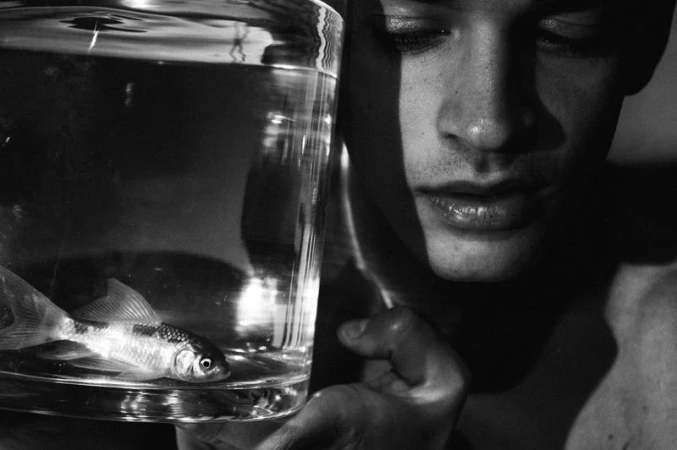 Black and white artistic portrait of a bare-chested man with a fish in a glass container, creating a conceptual and dramatic visual.