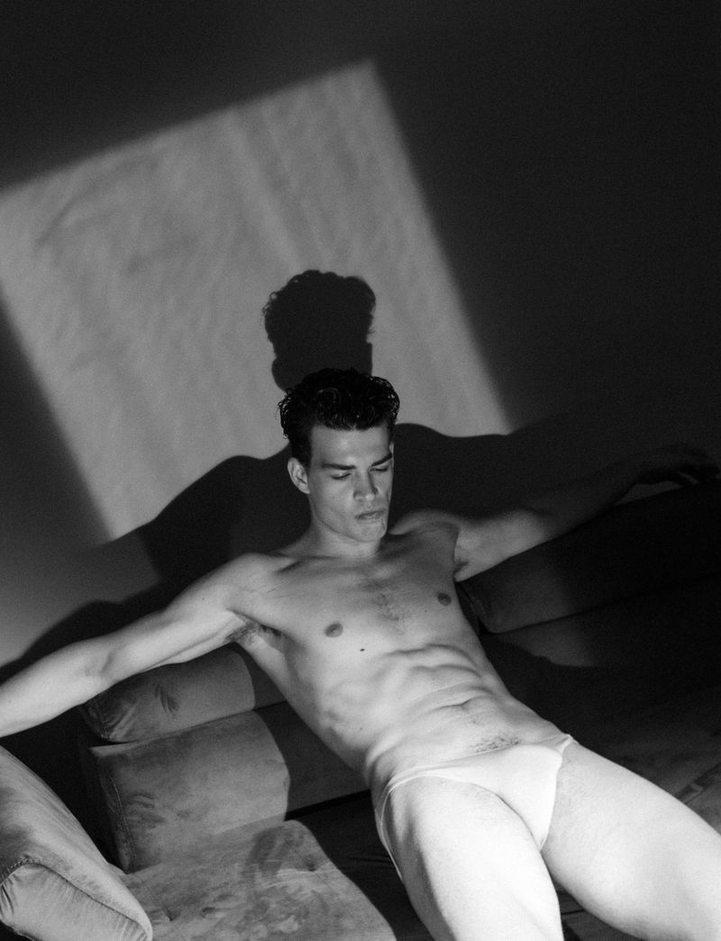Black and white portrait of a shirtless man reclining on a cushion in a photo studio, with dramatic shadow and light.