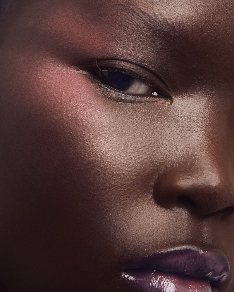 Close-up portrait of a dark-skinned woman with glossy lips, pink blush, and dramatic eye makeup, showcasing beauty and skincare.