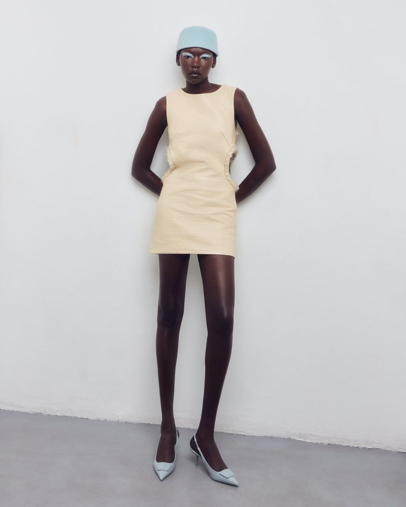 Model in cream mini dress wearing a light blue cap and pointed heels against a white seamless background