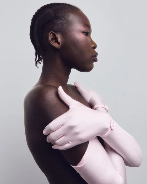 Striking portrait of a topless African woman with flawless skin, wearing elbow-length light pink gloves, looking off to the side in a studio.