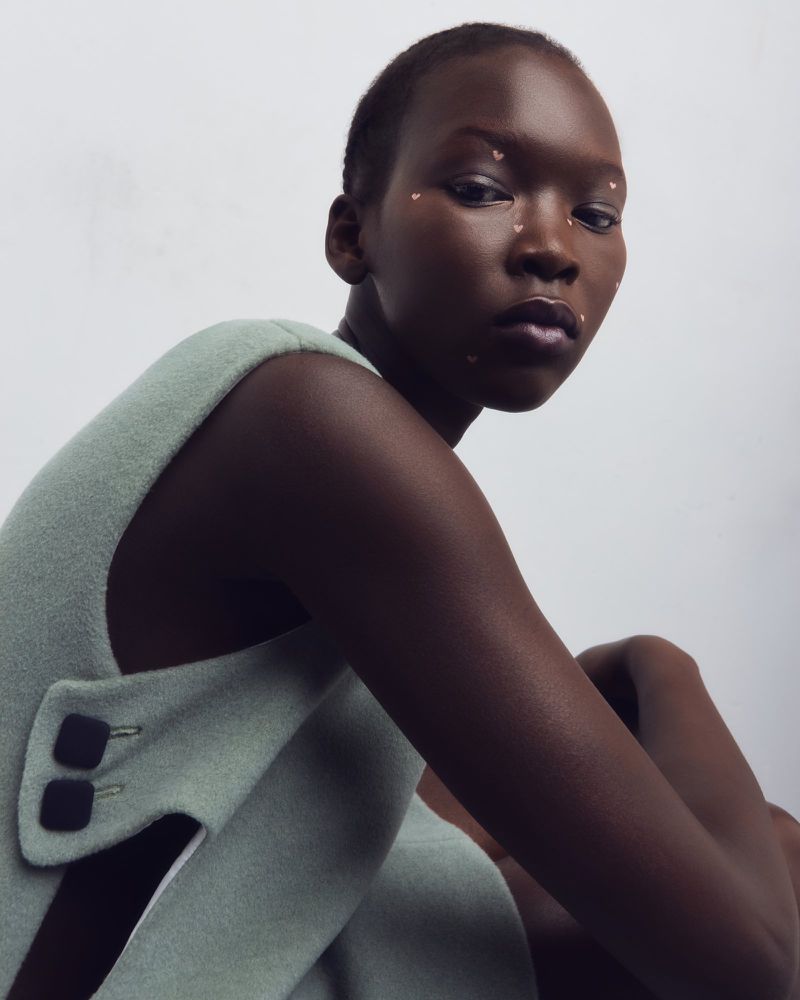 Portrait of a young Black woman with decorative facial cosmetic dots, posing in a light green wool dress against a white background.