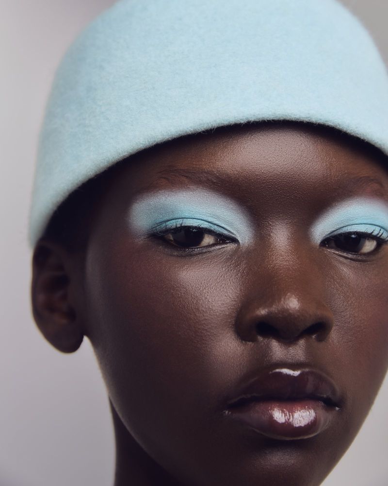 Close-up beauty shot of a dark-skinned woman with blue eyeshadow and a light blue hat against a neutral background. Glossy lips.