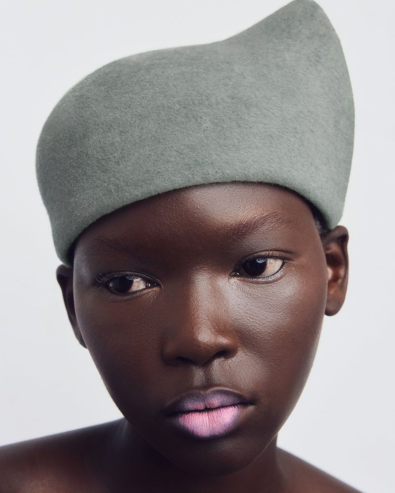 Close-up portrait of a dark-skinned woman with soft pink lipstick, wearing a soft sage-green felt hat against a plain white background.