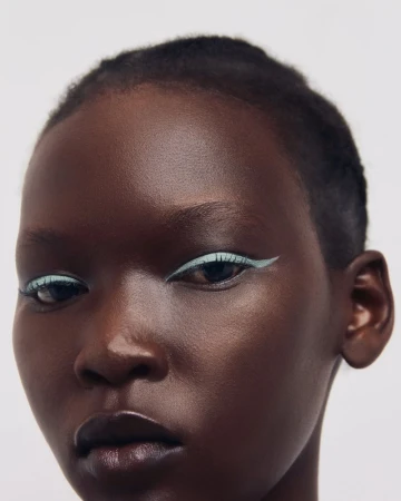 Close-up portrait of a model with short dark hair wearing light blue graphic eyeliner against a plain background