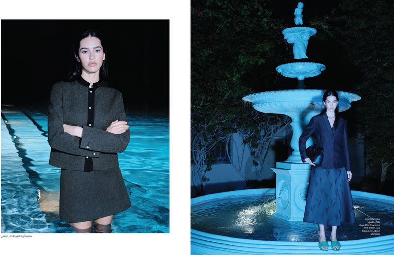 A fashion shot in a dark outdoor setting features an adult woman in a skirt suit beside a fountain and a pool. Low lighting creates a moody atmosphere.