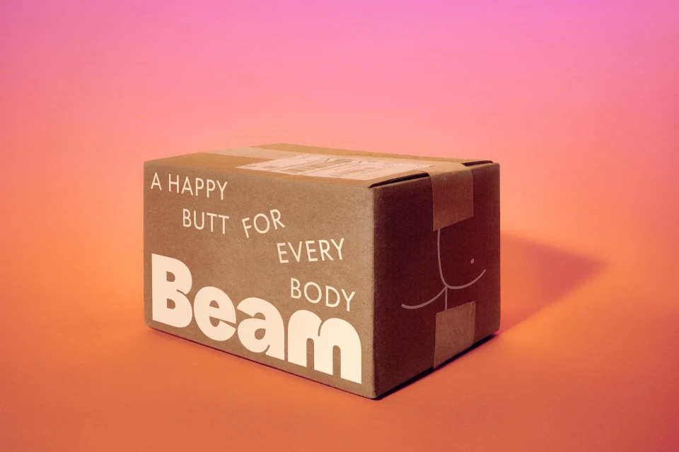 Brown cardboard box with white text 'A happy butt for every body' and a cheeky illustration, against a vibrant orange-to-pink gradient.