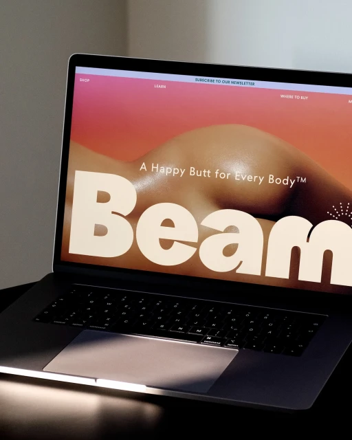 Laptop displaying the Beam website featuring a photo of a woman's bare back and buttocks. Text reads: "A Happy Butt for Every Body."