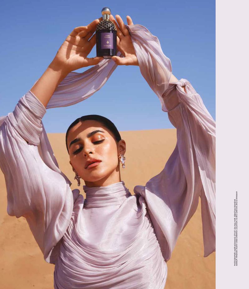 A woman in a lilac dress in the desert holds a perfume bottle against a blue sky, creating a serene and elegant beauty shot.