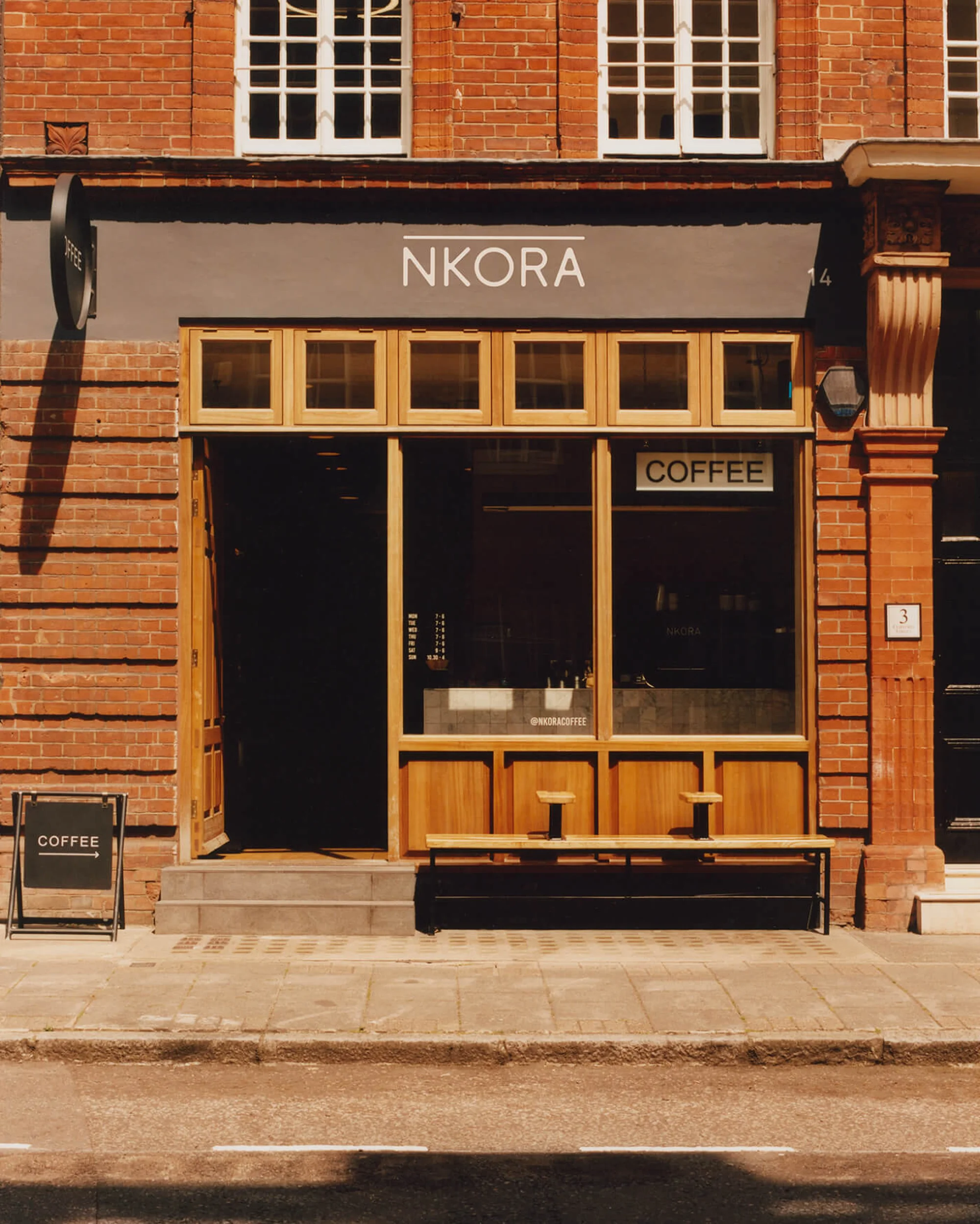 Exterior of NKORA coffee shop in brick building with wood frame windows, sidewalk, and street view on a sunny day.