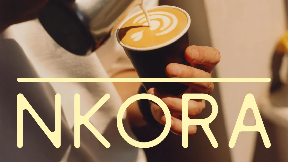 Person pours latte art into a cup of coffee.  Close-up of latte with coffee and white foam design on top with the text NKORA across frame.