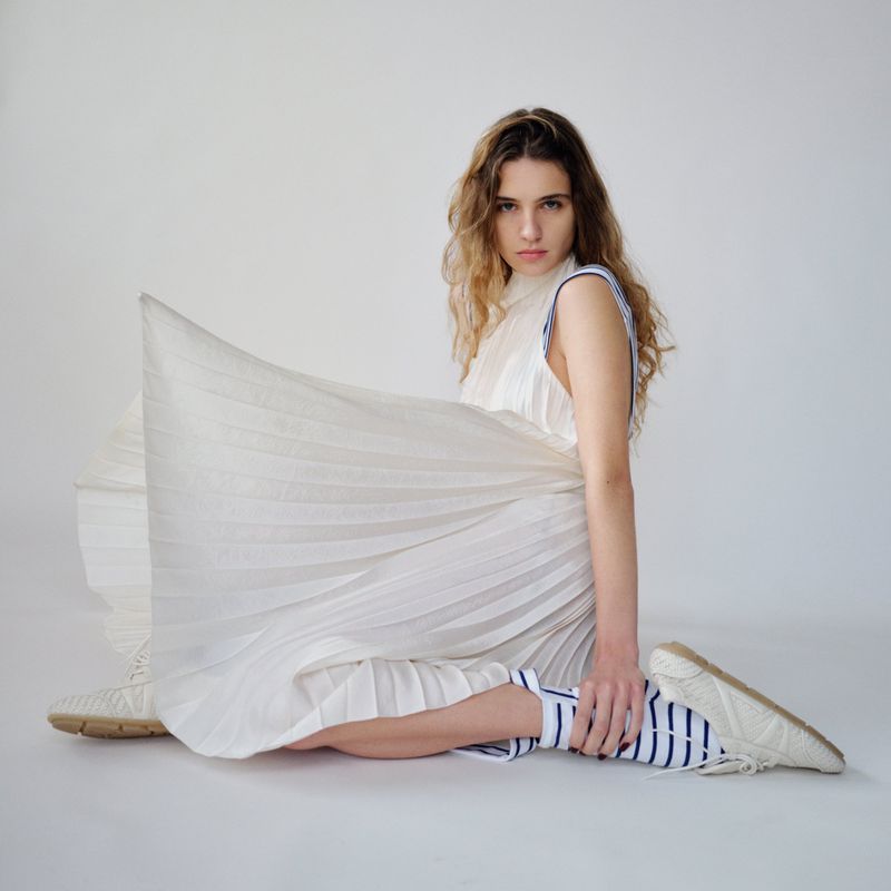 Model wearing white pleated dress and striped socks seated against white background for Financial Times HTSI Magazine