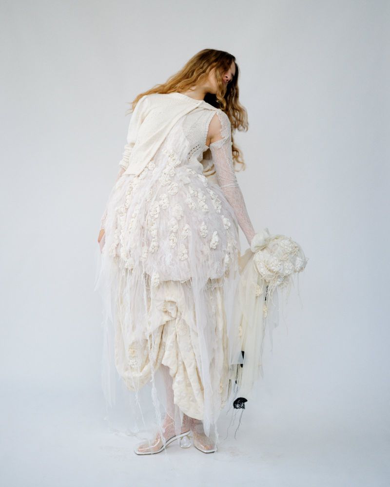 Full-length view of a slender woman in a textured ivory dress with tulle, sheer gloves, and heeled shoes, set against a plain white backdrop.