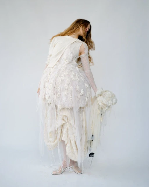 Full-length view of a slender woman in a textured ivory dress with tulle, sheer gloves, and heeled shoes, set against a plain white backdrop.