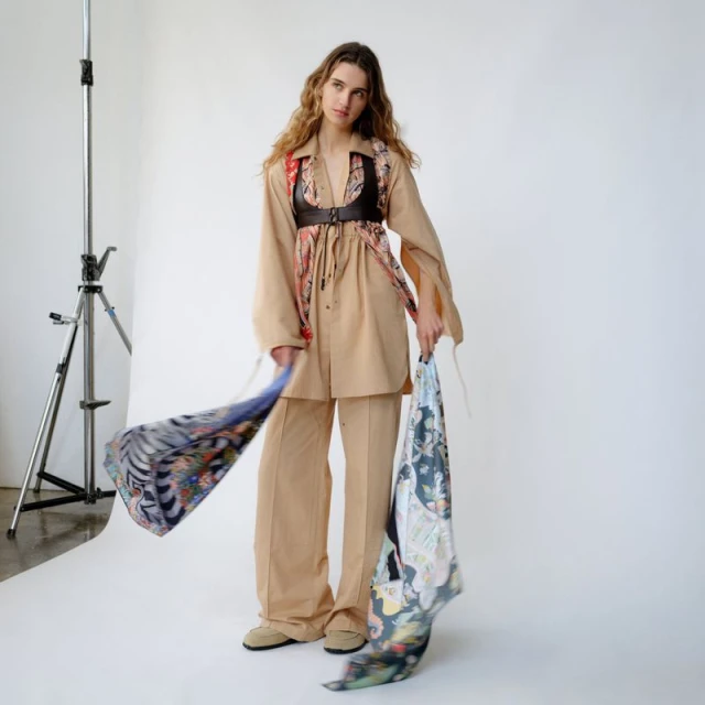 A long-haired fashion model in a tan outfit poses in a photo studio, holding a patterned scarf, with photo equipment visible in the background.