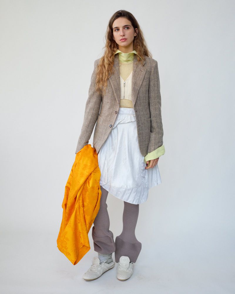 A woman in a blazer, skirt, and tights holds an orange garment in a studio setting against a white background in a fashion shot.