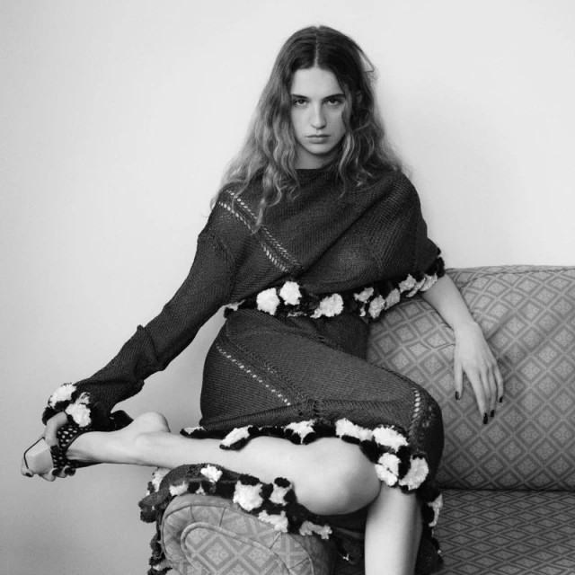 Monochrome portrait of a slender, fair-skinned woman with long hair sitting on an ornate sofa, wearing a knitted pompom dress.