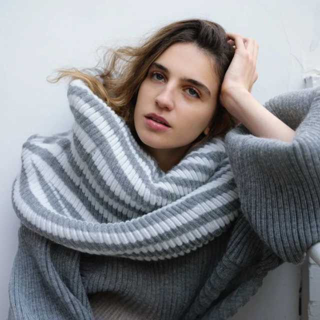 Woman in oversized grey knit sweater and striped scarf looking off-camera with hand on head, against neutral wall indoors.