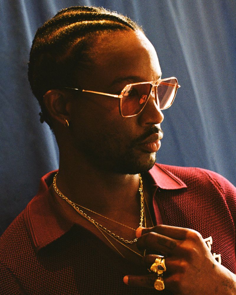 Portrait of a stylish African-American man with braided hair, wearing sunglasses and gold jewelry, posing against a blue background.