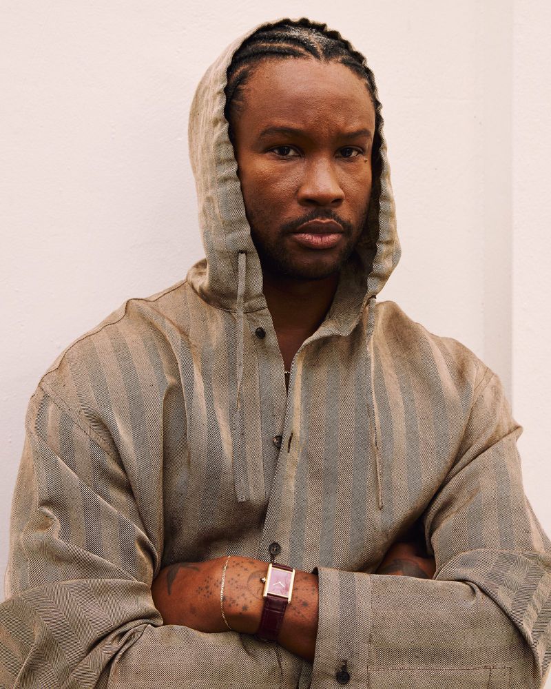 A man poses indoors with his arms crossed, wearing a hooded jacket, a wristwatch, and his hair in braids against a neutral backdrop.