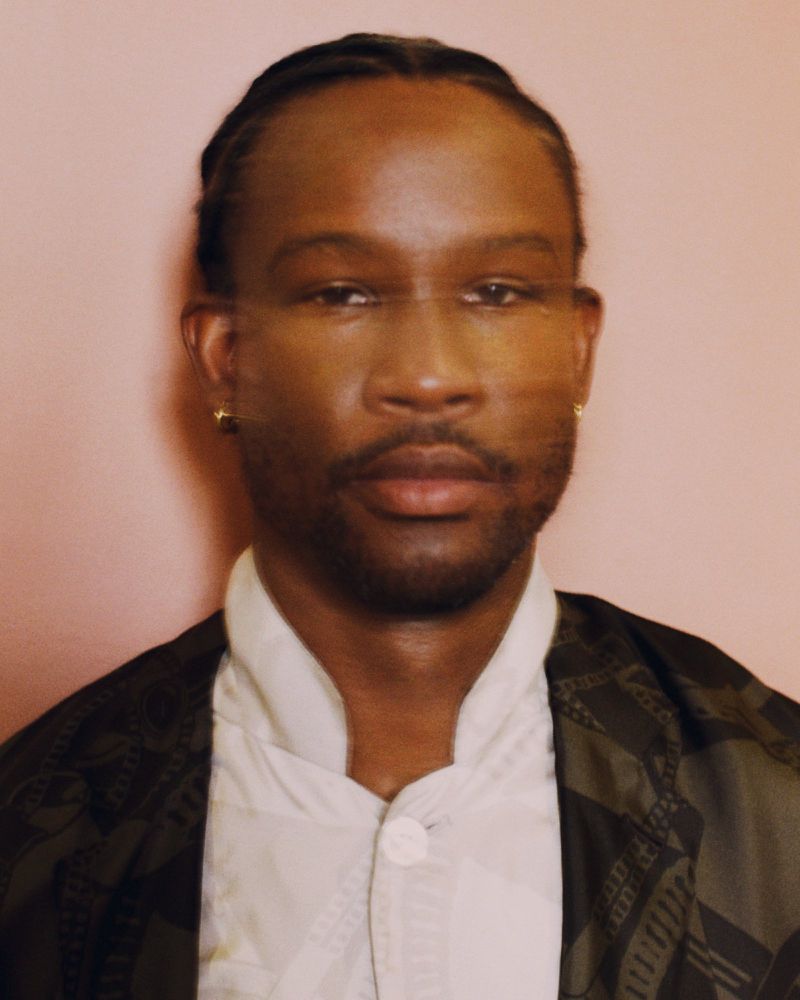 Portrait of a Black adult male wearing a white button-down shirt and patterned coat against a pale pink studio backdrop.