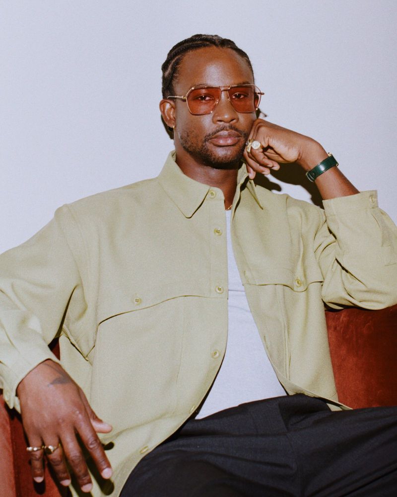 Portrait of a Black man in sunglasses and a light brown collared shirt, seated on a rust-colored couch against a light gray background.