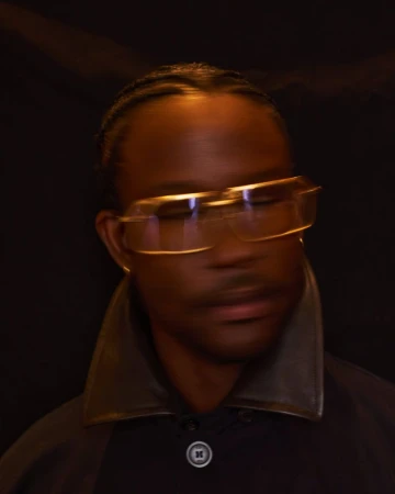 Portrait of a Black man wearing clear-framed glasses and a dark high-collared jacket against a dark background