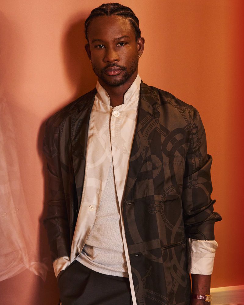 Portrait of Akinola Davies Jr in patterned dark blazer and light patterned shirt against a warm peach wall for Wonderland Magazine