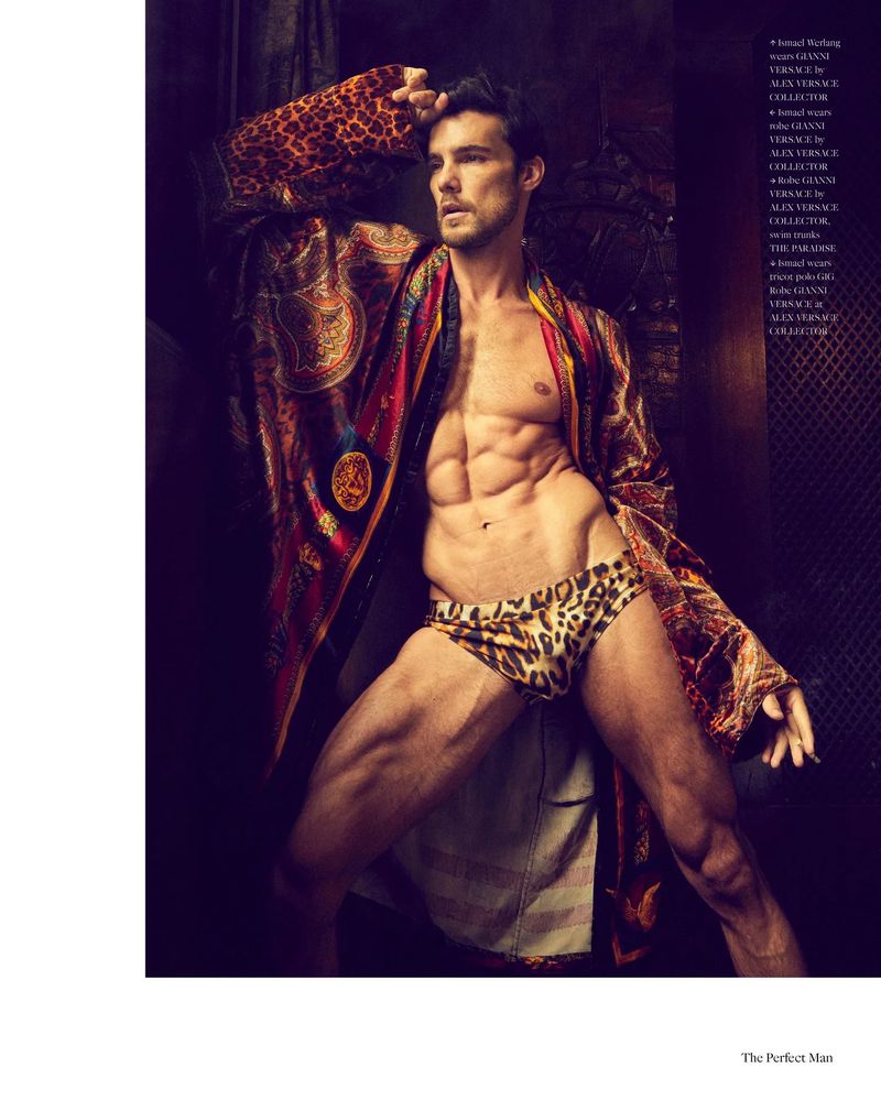 Fashion shot of a muscular adult male wearing leopard-print swimwear and patterned robe, posing indoors with strong lighting and shadows.